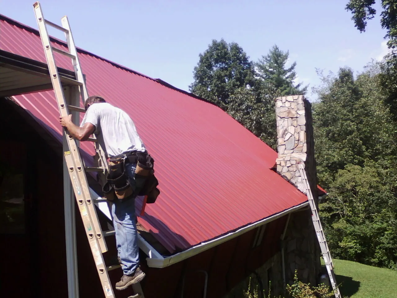 Roofer on a ladder beside a red metal roof and stone chimney — Soffit & Fascia Repair by Roofing Brothers in Millersport